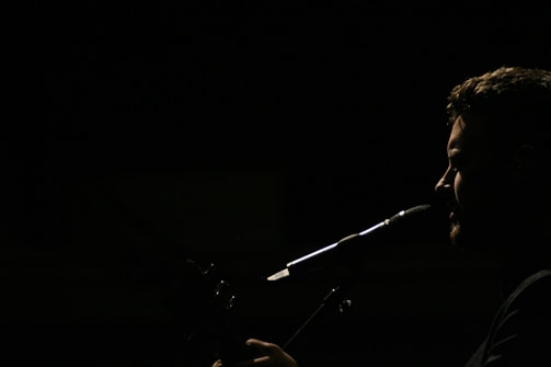 A silhouette of a person is captured in profile against a dark background, with light emphasizing their facial features and a microphone positioned near their mouth, suggesting they are singing or speaking.