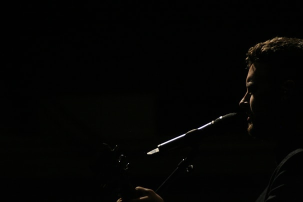A silhouette of a person is captured in profile against a dark background, with light emphasizing their facial features and a microphone positioned near their mouth, suggesting they are singing or speaking.