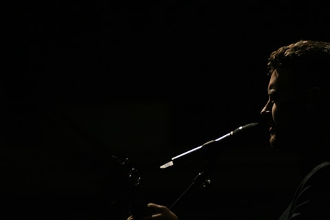A silhouette of a person is captured in profile against a dark background, with light emphasizing their facial features and a microphone positioned near their mouth, suggesting they are singing or speaking.