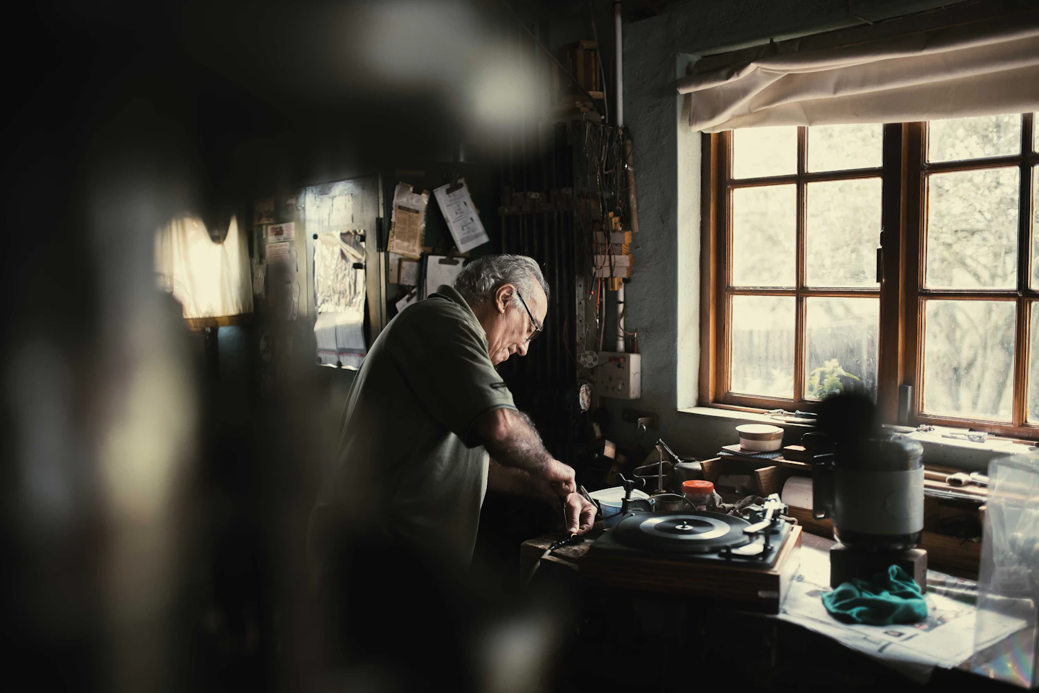 A man bent over a cluttered workbench under a single lamp, a dismantled record player in front of him
