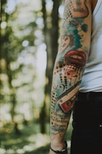 An arm with colorful tattoos in a forest setting. The tattoos include a variety of designs such as a heart with clock hands, a pair of lips with dripping ink, music symbols, and lettering. The tattoos are vibrant, featuring a mix of colors including blue, red, and black. The background consists of out-of-focus trees, creating a natural and serene environment.
