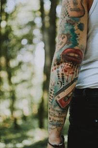 An arm with colorful tattoos in a forest setting. The tattoos include a variety of designs such as a heart with clock hands, a pair of lips with dripping ink, music symbols, and lettering. The tattoos are vibrant, featuring a mix of colors including blue, red, and black. The background consists of out-of-focus trees, creating a natural and serene environment.
