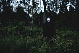 A person wearing a Guy Fawkes mask stands in a forest setting surrounded by dense trees and foliage. The atmosphere is dark and mysterious, with shadows dominating the scene.