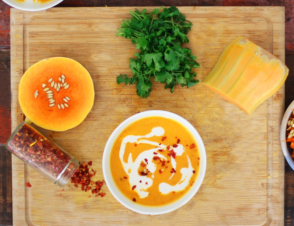 bowl of soup with two sliced vegetables, chili and green vegetable on brown wooden chopping board