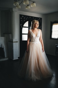 A woman in a luxurious, flowing bridal gown stands near a doorway, gazing thoughtfully into the distance. The dress features intricate lace detailing on the bodice and a voluminous tulle skirt. The room is softly lit, creating a serene and contemplative atmosphere, with vintage-style decor visible in the background.