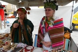 Two people are wearing traditional clothing with colorful patterns and headdresses. They are standing behind a market stall displaying various handmade crafts, such as beaded jewelry and woven textiles. The setting appears to be outdoors, with a white tent and a street view in the background.