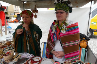 Two people are wearing traditional clothing with colorful patterns and headdresses. They are standing behind a market stall displaying various handmade crafts, such as beaded jewelry and woven textiles. The setting appears to be outdoors, with a white tent and a street view in the background.
