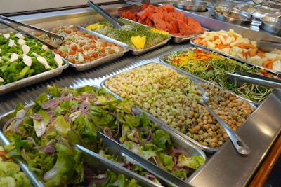 An assortment of fresh and colorful salad ingredients are displayed in metal trays. The variety includes greens, chickpeas, sliced onion, cherry tomatoes, shredded carrots, sprouts, and watermelon slices. Tongs are placed in several trays for serving.