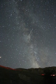 A star-filled night sky with the Milky Way visible in the center, set above a silhouette of mountainous terrain. A bright meteor streaks across the sky.