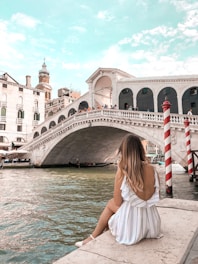 A woman in a white dress with stripes is sitting on the edge of a canal, facing away from the camera and looking towards a large arched stone bridge. The bridge is the famous Rialto Bridge in Venice, with several people standing on it. Venetian architectural buildings are visible in the background. The sky is partly cloudy. There are red and white striped poles in the water, which is typical for Venice.