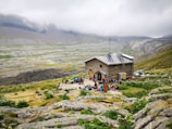 A rustic stone building is situated in a mountainous landscape, surrounded by green fields and rocky terrain. A group of people is gathered in front of the building, engaging in outdoor activities. The sky is overcast with low-hanging clouds enveloping the distant mountains.