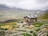 A rustic stone building is situated in a mountainous landscape, surrounded by green fields and rocky terrain. A group of people is gathered in front of the building, engaging in outdoor activities. The sky is overcast with low-hanging clouds enveloping the distant mountains.