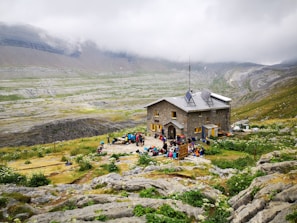 A rustic stone building is situated in a mountainous landscape, surrounded by green fields and rocky terrain. A group of people is gathered in front of the building, engaging in outdoor activities. The sky is overcast with low-hanging clouds enveloping the distant mountains.
