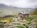 A rustic stone building is situated in a mountainous landscape, surrounded by green fields and rocky terrain. A group of people is gathered in front of the building, engaging in outdoor activities. The sky is overcast with low-hanging clouds enveloping the distant mountains.