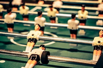 Several foosball player figures attached to rods are positioned on a green foosball table. The figures are wearing white shirts and black shorts, with the focus on one figure in the foreground.