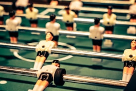Several foosball player figures attached to rods are positioned on a green foosball table. The figures are wearing white shirts and black shorts, with the focus on one figure in the foreground.