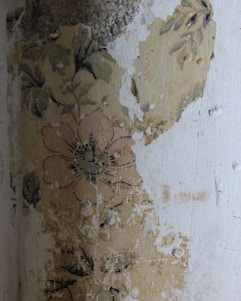 A section of an old wall with peeling paint and faded floral wallpaper, showcasing a large flower and leaves. The surface appears worn and distressed, indicating age and weathering.