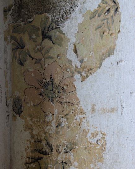 A section of an old wall with peeling paint and faded floral wallpaper, showcasing a large flower and leaves. The surface appears worn and distressed, indicating age and weathering.