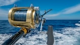 A close-up view of a fishing rod and reel on a boat, angling out over a deep blue ocean with a clear sky and a few clouds. The reel is shiny, metallic, and well-crafted, indicating it is likely for deep-sea fishing.