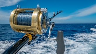 A close-up view of a fishing rod and reel on a boat, angling out over a deep blue ocean with a clear sky and a few clouds. The reel is shiny, metallic, and well-crafted, indicating it is likely for deep-sea fishing.