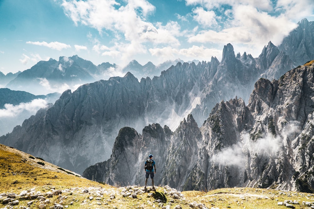 Person, die auf einem grüngrasigen Berg mit Blick auf graue felsige Berge steht, die tagsüber von Wolken bedeckt sind