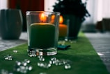 A lit candle in a clear glass holder is placed on a green tablecloth, surrounded by scattered decorative glass beads. Another candle and a plant are blurred in the background, creating a cozy and serene ambiance.