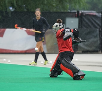A field hockey goalkeeper in red protective gear is actively blocking a ball on an artificial green turf field. In the background, another player dressed in black holds a hockey stick and is out of focus.