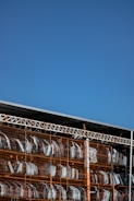 A large outdoor storage area with metal shelves containing multiple car bumpers, arranged in rows and columns. The sky is clear and blue, forming the backdrop of the image.