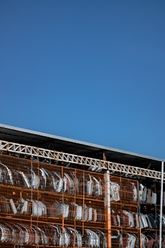 A large outdoor storage area with metal shelves containing multiple car bumpers, arranged in rows and columns. The sky is clear and blue, forming the backdrop of the image.