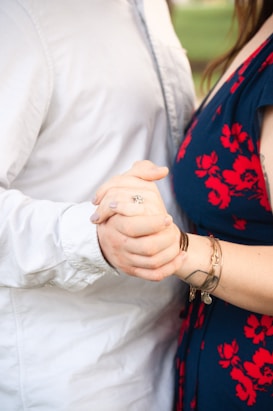 A close-up of two people holding hands, one wearing a white shirt and the other wearing a dark blue dress with red floral patterns. The individuals are connected in a gentle and intimate manner, with one person showing a silver ring on their finger.
