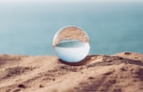 A crystal ball rests on a sandy surface, reflecting an inverted image of the ocean and sky behind it. The background is softly blurred, emphasizing the clear sphere and the serene seascape within it.