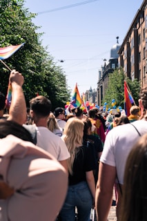 A crowd of people is gathered outdoors, surrounded by buildings and trees. Many are holding or wearing rainbow flags, indicative of a pride event or celebration. Brightly colored balloons can be seen above the crowd, and the sky is clear and sunny.