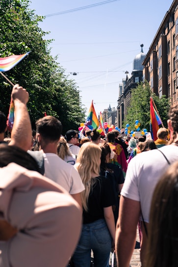 A crowd of people is gathered outdoors, surrounded by buildings and trees. Many are holding or wearing rainbow flags, indicative of a pride event or celebration. Brightly colored balloons can be seen above the crowd, and the sky is clear and sunny.