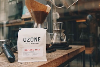 A bag of Ozone Coffee Roasters, labeled 'Gaharo Hill,' is placed on a wooden surface. The bag is white with black and red text. Behind it, there are various coffee-making tools, including a pour-over coffee dripper with a filter, carafes, and a tamper. The setting appears to be in a cozy coffee shop or a barista station.