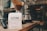 A bag of Ozone Coffee Roasters, labeled 'Gaharo Hill,' is placed on a wooden surface. The bag is white with black and red text. Behind it, there are various coffee-making tools, including a pour-over coffee dripper with a filter, carafes, and a tamper. The setting appears to be in a cozy coffee shop or a barista station.