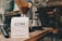 A bag of Ozone Coffee Roasters, labeled 'Gaharo Hill,' is placed on a wooden surface. The bag is white with black and red text. Behind it, there are various coffee-making tools, including a pour-over coffee dripper with a filter, carafes, and a tamper. The setting appears to be in a cozy coffee shop or a barista station.