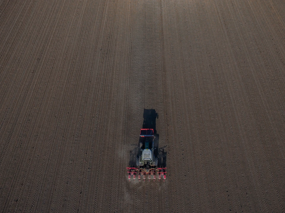 The image presents an aerial view of a large, flat field with a tractor at its center. The tractor is actively engaged in plowing the field, creating furrows that will be used for planting crops. The tractor's path across the field is clearly visible, indicating it is moving from left to right and slightly above the ground level.