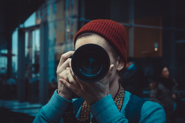 A person in a red beanie is holding a camera with a large lens up to their face, partially obscuring it. The background consists of urban architecture with reflective glass walls, and some people are visible in the distance.