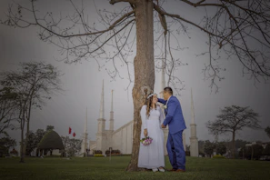 A couple, dressed in formal wedding attire with the bride holding a bouquet, stand closely by a large tree. The groom wears a blue suit while the bride is in a white dress adorned with a floral headband. In the background are tall spires of a church-like building and well-manicured gardens, along with two red flags.