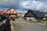 Charming rural scene with several houses, some traditional and made of wood, showing a blend of modern and classic architectural styles. A paved road curves around the houses, surrounded by a wooden fence and some greenery. The sky above is partly cloudy, casting soft shadows on the structures.