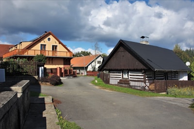 Charming rural scene with several houses, some traditional and made of wood, showing a blend of modern and classic architectural styles. A paved road curves around the houses, surrounded by a wooden fence and some greenery. The sky above is partly cloudy, casting soft shadows on the structures.