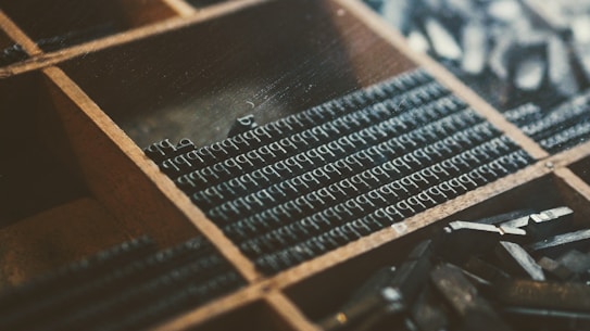 A close-up view of a wooden typesetting tray with small metal letter blocks organized in compartments. The arrangement suggests an old-fashioned printing technique, where each block represents an individual letter, likely for letterpress printing. The color palette has a vintage, muted tone, emphasizing shades of dark brown and deep grey.