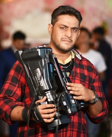 A person wearing a red and black checkered shirt is holding a large video camera. The individual appears to be focused and is set against a blurred background with subtly visible people.