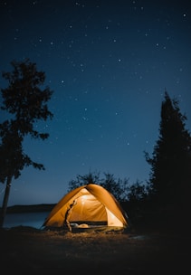 A lit-up camping tent is set amidst a natural landscape under a clear, starry night sky. There are silhouetted trees surrounding the tent, which is positioned near a body of water, creating a serene and peaceful atmosphere.