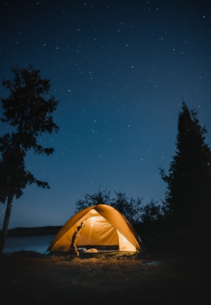 A lit-up camping tent is set amidst a natural landscape under a clear, starry night sky. There are silhouetted trees surrounding the tent, which is positioned near a body of water, creating a serene and peaceful atmosphere.