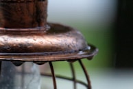Close-up view of an aged, textured copper object with droplets of water on its surface. The copper appears weathered, with subtle curves and patina, capturing small details and reflections. The background is blurred, with hints of green and brown, suggesting an outdoor setting.