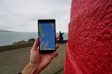 A hand is holding a smartphone displaying a map application. Red brick wall on the right side and a person in the background near a body of water.