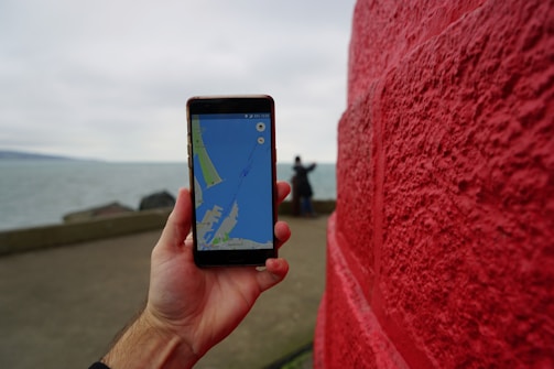 A hand is holding a smartphone displaying a map application. Red brick wall on the right side and a person in the background near a body of water.