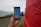 A hand is holding a smartphone displaying a map application. Red brick wall on the right side and a person in the background near a body of water.