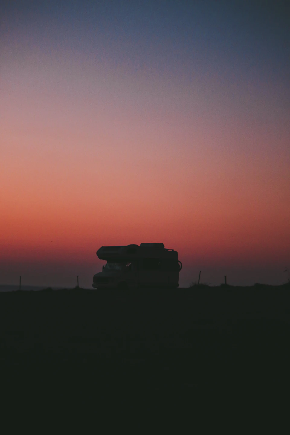The image depicts a white RV parked on a grassy field, facing towards the right side of the frame and set against an orange sky with a pinkish hue. The scene is bathed in soft light, creating a serene atmosphere. In the distance, there are trees visible, adding depth to the landscape.